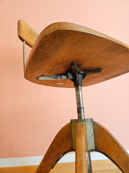 Girsberger Stuhl Ein vintage Holzstuhl mit verstellbarem Fuß und grauer Wand im Hintergrund.