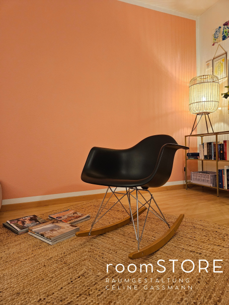Vitra Rocking Armchair Rod Base 420.- Schwarzer Schaukelstuhl vor einem orangefarbenen Wand, mit Zeitschriften auf dem Boden.